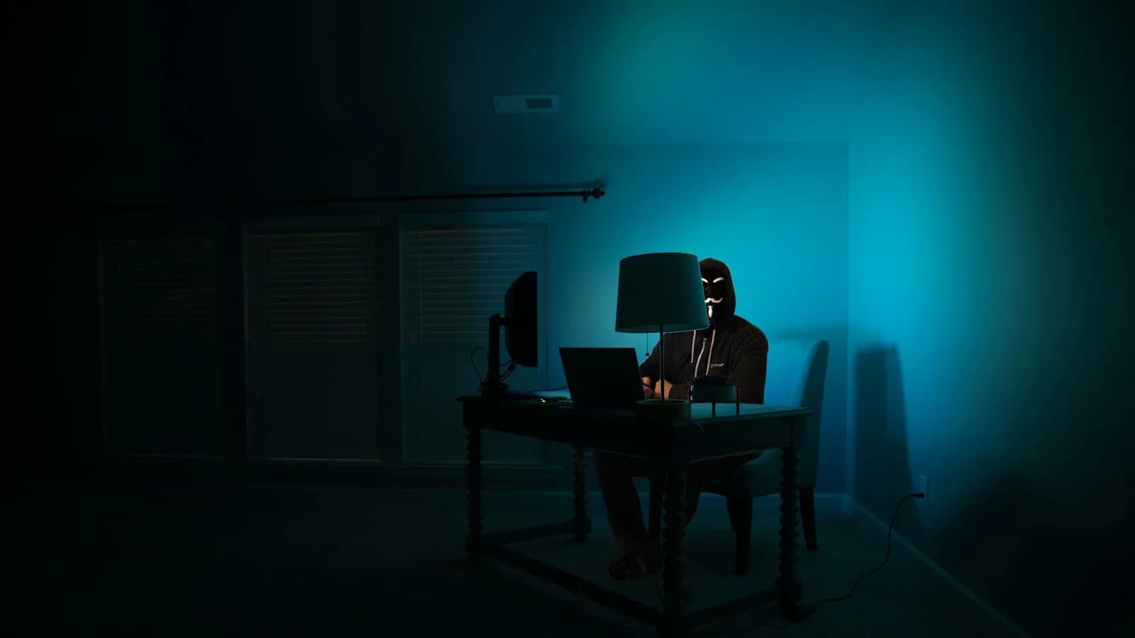 A person in a hood working on a laptop in a dark room lit in teal